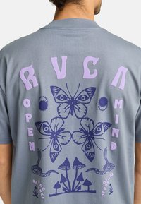 Camiseta de algodón gris con un diseño gráfico en púrpura de mariposas, serpientes y hongos, con el texto "MENTE ABIERTA" y "RVCA."