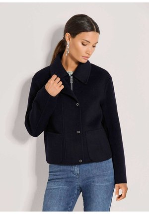 Frau trägt eine schwarze Jacke mit Reißverschluss und Knöpfen mit großem Kragen, kombiniert mit blauen Jeans, schaut nach unten und berührt die Jacke am Kragen.