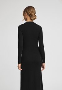 Svart ribbad stickad klänning med långa ärmar, figursydd silhuett och hög krage. Slät textur och golvlång design. Inga synliga mönster eller accenter.