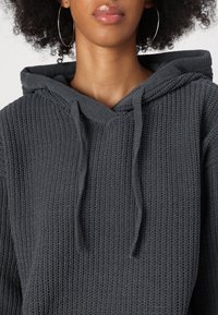 Grijze gebreide hoodie met een gemêleerd patroon, V-hals en koorden aan de capuchon. Dikke stof, zichtbare naden en een losse pasvorm.