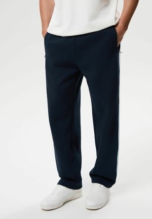 Person trägt dunkelblaue Hose mit Seitentaschen und weiße Sneakers, steht mit Händen in den Taschen vor einem einfarbigen Hintergrund.