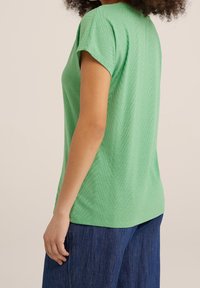 Žena v krátkém rukávu zeleném texturovaném topu a tmavě modrých kalhotách, zobrazená z boku na jednolitém pozadí.