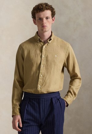 Koszula khaki na guziki wykonana z tkaniny o teksturze, z klasycznym kołnierzem i małym logo na piersi, noszona z granatowymi spodniami w paski.