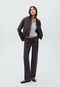 Kvinna klädd i en svart kort jacka, ljusgrå ribbad topp, svarta jeans med rhinestones på fickorna och svarta platta skor som står mot en enfärgad bakgrund.