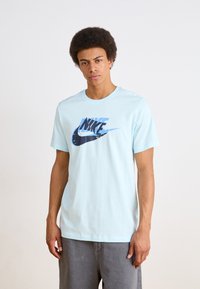 Světle modré bavlněné tričko s tmavě modrým logem Nike a grafickým designem. Klasický výstřih, krátké rukávy, uvolněný střih.