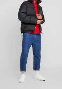 Czarna kurtka puchowa z wysokim kołnierzem, czerwona koszula pod spodem, niebieskie dżinsy i białe sneakersy. Gładka faktura, luźny krój.