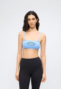 Νεαρή γυναίκα με σγουρά σκουρόχρωμα μαλλιά, φορώντας ένα ανοιχτό μπλε crop top Calvin Klein και ψηλόμεσα μαύρα κολάν, στέκεται μπροστά σε απλό φόντο.