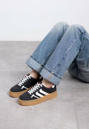 Zapatillas negras con rayas blancas y acentos rojos, con cordones planos y suela de goma beige. Llevadas con jeans de mezclilla azul claro doblados.
