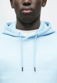 Gaiši zils džemperis ar vaļīgu piegriezumu, auklām un maigu tekstūru. Apkakles zonā redzama balta iekšējā kārta.