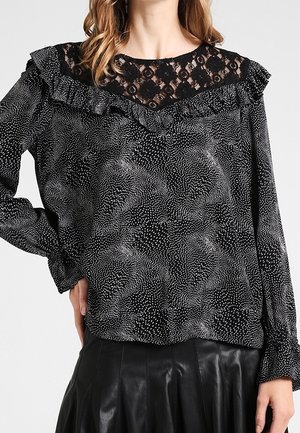 Schwarze Bluse mit einem Spitzenbesatz und gerüschtem Ausschnitt, ausgestattet mit einem strukturierten Muster aus weißen Punkten; kombiniert mit einem schwarzen, gefalteten Lederrock.