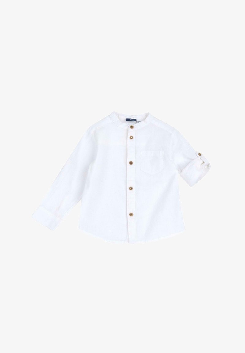 Camicia bianca a maniche lunghe in tessuto leggero con colletto rotondo, chiusura frontale con bottoni e taschino sul petto. I polsini abbottonati sono arrotolati.