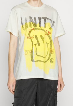 Person trägt weißes T-Shirt mit großem gelbem Smiley-Gesicht, Schriftzug "UNITY", roter Kronen-Kritzelei und schwarze Cargo-Hose.