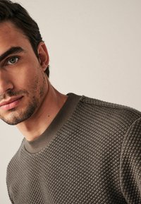 Uomo con capelli corti e scuri e leggera peluria che indossa una camicia a maniche lunghe marrone texture su uno sfondo beige semplice.