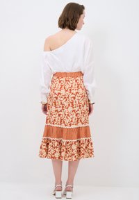 Weiße Bluse mit schulterfreiem Design, kombiniert mit einem gemusterten orangefarbenen Rock, der Blumenmuster und einen rüschierten Saum aufweist.