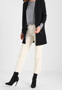 Cardigan nero testurizzato, top grigio chiaro, jeans crop color crema e stivali neri con tacco alto. Dettagliato con orli sfilacciati, silhouette aderente.