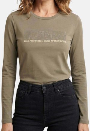 Donna che indossa una maglia a maniche lunghe verde oliva con testo in strass che recita "FRIDAY" e slogan "Less perfection more authenticity,", abbinata a jeans neri.
