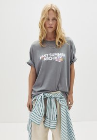 Blond kvinde med bølget hår iført grå t-shirt med rullede ærmer med teksten "Past Summer Archives", stribet skjorte bundet om taljen og lyse bukser.