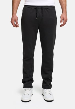 Homme portant un pantalon de survêtement noir à cordon et des baskets blanches, debout devant un fond clair uni.