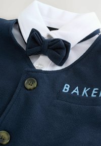 Tumši zils bērnu krekls ar baltu apakšmalu, teksturēts audums, ar tauriņa kaklasaiti un izšūtu "BAKER" uz kabatas. Piedāvā divas pogas.
