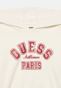 Φούτερ με κουκούλα σε κρεμ χρώμα, με έντονα κόκκινα γράμματα που αναγράφουν "GUESS PARIS". Λείο ύφασμα με μπροστινή τσέπη καγκουρό και κουκούλα.