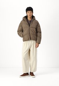 Brown puffer jacket with a hood, zip closure, and stitched sections; paired with loose beige trousers and brown sneakers.