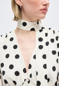 Γυναίκα που φοράει λευκό μπλουζάκι με μεγάλα μαύρα πουά και ταιριαστό μαντήλι στον λαιμό, παρουσιάζοντας το πλάι του προφίλ της και σκουλαρίκια.