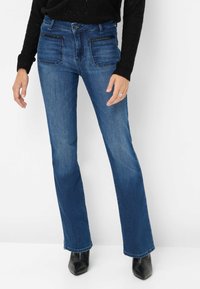 Jeans a zampa d'elefante in denim blu con tasche anteriori e posteriori, caratterizzati da dettagli neri lungo le tasche. Indossati con stivali alla caviglia neri con punta a punta.