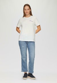 White cotton t-shirt with short sleeves, featuring blue embroidered text. Paired with light blue distressed jeans and black sneakers.