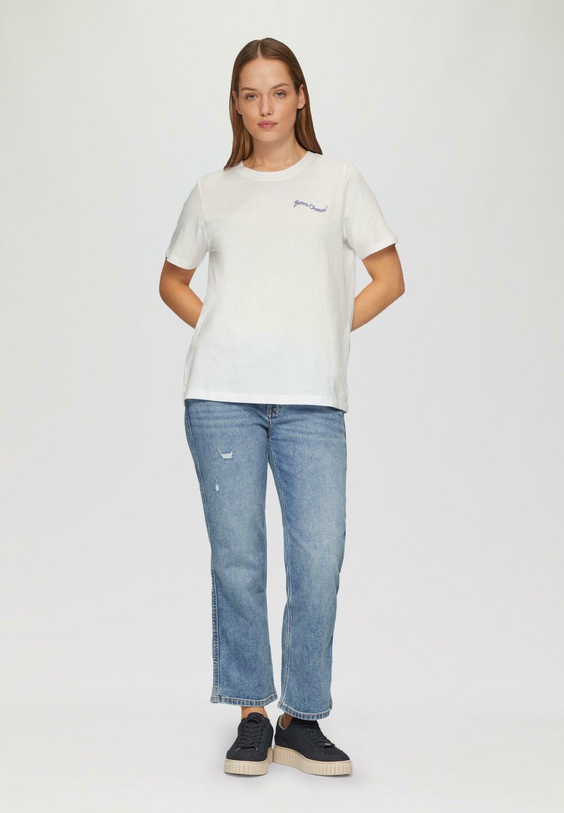 White cotton t-shirt with short sleeves, featuring blue embroidered text. Paired with light blue distressed jeans and black sneakers.