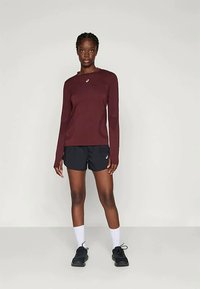 Žena stoji u crnim šortsevima, bordo dugim rukavima, bijelim čarapama i crnim patikama na jednostavnoj svijetloj pozadini.