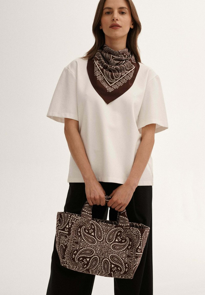 Kobieta w białej bluzce z krótkim rękawem oraz brązowo-białym bandamą w wzór paisley na szyi, trzymająca torebkę w pasującym wzorze.