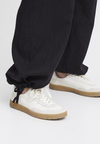Baskets en cuir blanc avec une semelle en caoutchouc, présentant des détails en dentelle et un embout texturé, associées à un pantalon noir à poignets élastiques froncés.