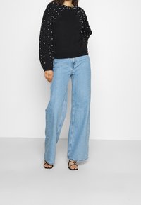 Maglione nero a maglia con dettagli in perle sulle maniche, abbinato a jeans larghi azzurri e sandali neri con cinturini. Sfondo semplice.