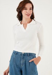 White ribbed long-sleeve polo shirt with a v-neck collar, paired with high-waisted blue denim jeans; hands in pocket.