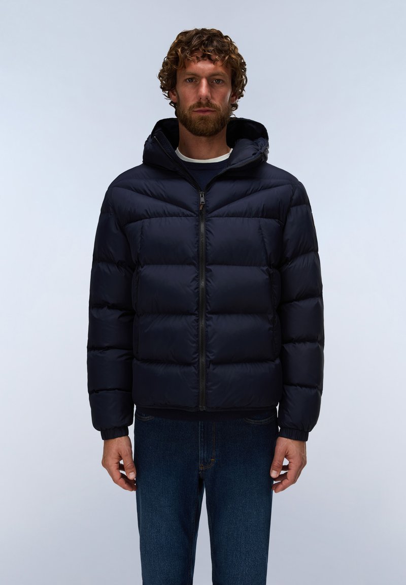 Man met krullend haar en baard, gekleed in een zwarte gewatteerde jas met rits en capuchon en blauwe jeans tegen een effen lichte achtergrond.