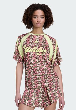 Ung kvinde med krøllet hår iført matchende blomsterprintet Adidas t-shirt og shorts i lyserøde, grønne og creme farver, stående foran en ensfarvet baggrund.