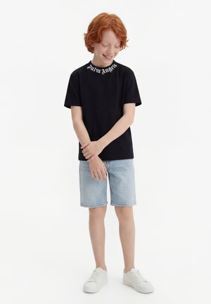 Ragazzo dai capelli rossi che indossa una t-shirt nera Palm Angels, pantaloncini di jeans azzurri e sneaker bianche, sorridente con le braccia incrociate davanti.