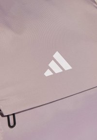 Mochila de color púrpura claro con cierre plano, herrajes negros y un logo blanco de tres rayas en el frente. Superficie de tela texturizada.