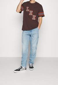 Camiseta marrón con rayas rojas y azules en las mangas, con un estampado que dice "LEE". Pantalones vaqueros azul claro, zapatillas negras con cuadros.