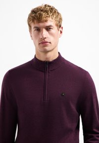 Pullover-Sweater aus tiefviolettem Strickstoff mit hohem Kragen und Reißverschluss. Besitzt ein kleines schwarzes sechseckiges Logo auf der linken Seite.