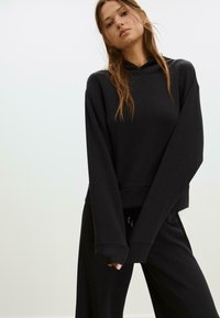 Młoda kobieta z długimi brązowymi włosami, ubrana w oversize'owy czarny hoodie oraz pasujące szerokie spodnie, na jednolitym tle.