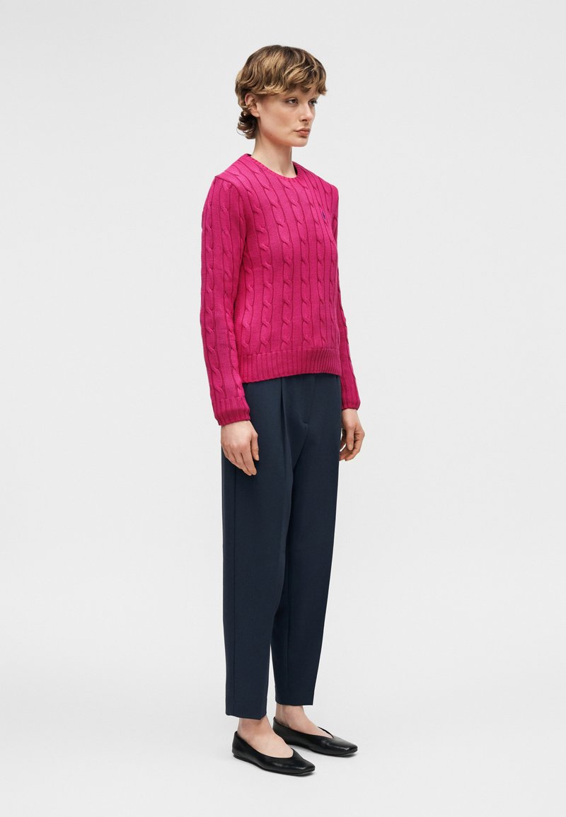 Femme debout de profil portant un pull tricoté torsadé rose vif, un pantalon foncé ajusté et des ballerines noires, aux cheveux courts et bouclés.