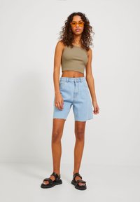 Top scurt de culoare verde măsliniu, pantaloni scurți din denim bleu deschis cu talie înaltă și margini desfăcute, asortați cu sandale negre cu talpă groasă și ochelari de soare portocalii.
