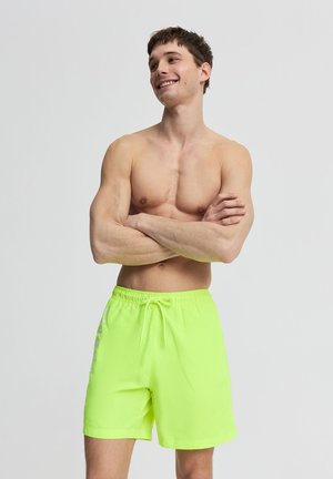 Giovane uomo muscoloso che sorride con le braccia incrociate, indossa pantaloncini da bagno verde neon brillante su uno sfondo chiaro e uniforme.