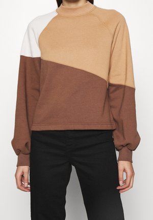 Kobieta nosząca longsleeve z blokami kolorów w odcieniach beżu, brązu i bieli, dobrany do czarnych spodni, widoczna od szyi do bioder.