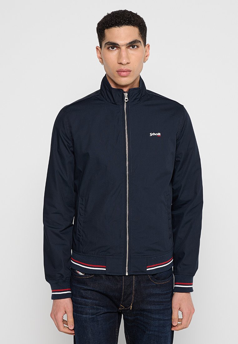 Veste bleu marine avec col montant, fermeture éclair à l'avant, accents rayés rouges et blancs aux poignets et à l'ourlet, fabriquée à partir d'un matériau léger et lisse.
