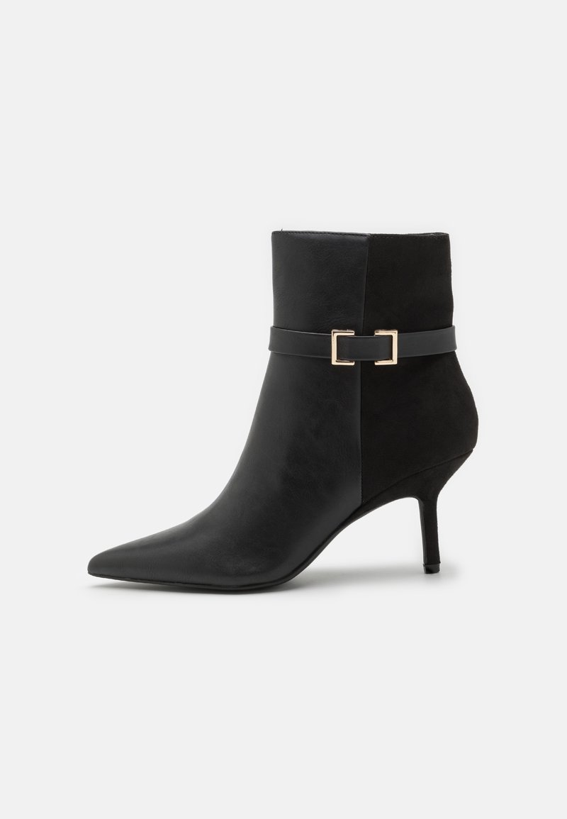 Bottine noire en cuir et daim, avec un bout pointu, un talon stiletto et une sangle décorative ornée d'une boucle dorée.