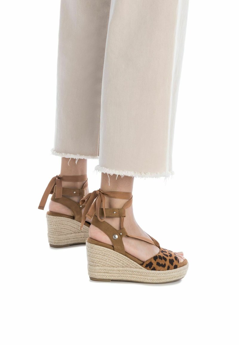 Sandalias de cuña marrones con tela de leopardo, lazos en los tobillos y suelas envolvidas en yute. Presentan un acabado texturizado y acentos decorativos de herrajes.