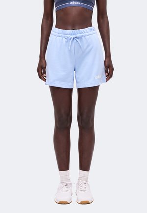 Γυναίκα με ανοιχτό μπλε σορτς Adidas, λευκές κάλτσες, λευκά αθλητικά παπούτσια και σκούρο μπλε αθλητικό μπουστάκι Adidas, όρθια μπροστά από μια απλή φόντο.
