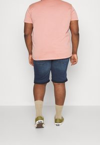 Camisa de manga corta rosa claro, pantalones cortos de mezclilla oscura, zapatos verde oliva y calcetines beige. Costuras visibles y dobladillos de los pantalones cortos enrollados.